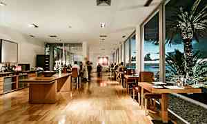 En restaurang med en modern interiör, träbord och stolar, samt stora fönster mot en tropisk utsikt med palmer och hav. (AI-genererad text)