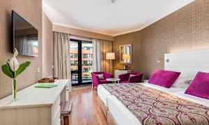 Hotellrum med dubbelsäng, skrivbord, två lila fåtöljer och balkong. (AI-genererad text)