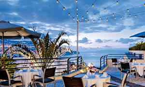 Uteservering med dukade bord och stolar, omgiven av ljusslingor och en utsikt över staden och havet. (AI-genererad text)