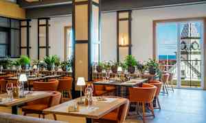 Ett välbelyst restauranginteriör med bord, stolar och grön växtdekoration. I bakgrunden finns stora fönster som visar en vy av en byggnad med ett klocktorn. (AI-genererad text)