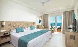 Ett hotellrum med dubbelsäng, sittgrupp och balkong med havsutsikt. (AI-genererad text)