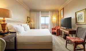 Ett hotellrum med en stor säng, nattduksbord, soffa, TV och ett skrivbord. Gardiner och glasdörrar leder till en balkong. (AI-genererad text)