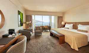 Hotellrum med dubbelsäng, sittgrupp och balkong med utsikt över havet. (AI-genererad text)