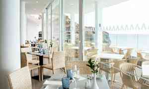 Ljust och öppet matområde med dukade bord. Utsikt över havet genom glasväggar. (AI-genererad text)