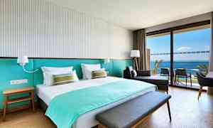 Ett hotellrum med en dubbelsäng, en sittmöbel och en balkong med utsikt över havet. (AI-genererad text)