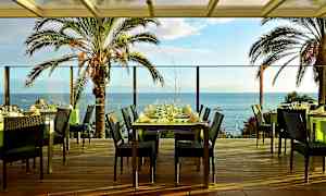 Restaurangbord på en terrass med utsikt över havet och palmer. (AI-genererad text)