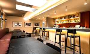 Ett modernt barområde med främst svarta bord och stolar samt en välbelyst bardisk fylld med flaskor och glas. (AI-genererad text)