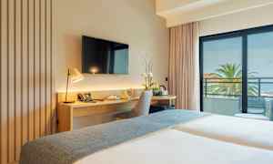 Ett hotellrum med skrivbord, stol och TV, samt en balkong med utsikt över palmer och havet. (AI-genererad text)