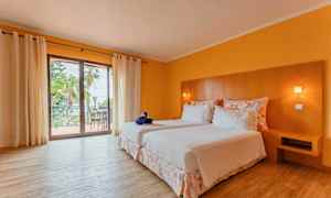 Ett hotellrum med två sängar klädda i vita sängkläder, ett trähuvudgavel och en balkong med utsikt. Rummet har gulmålade väggar och trägolv. (AI-genererad text)