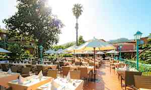 En utomhusterrass med uppdukade bord omgivna av parasoller och gröna växter. I bakgrunden syns en byggnad omgiven av träd. (AI-genererad text)