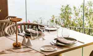 Ett dukat bord med glas och tallrikar, vid sidan av en glasvägg som vetter mot utomhus. (AI-genererad text)