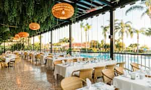 En ljus restaurang med dukade bord, omgiven av grönska med utsikt över en pool och palmträd. (AI-genererad text)