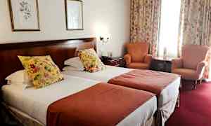 Ett hotellrum med två enkelsängar, dekorerade med blommiga kuddar och röda överkast. I rummet finns också två fåtöljer, ett litet bord och gardiner med blommönster. (AI-genererad text)