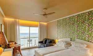 Ett hotellrum med två sängar, ett mönstrat grönt tapetfondvägg och en balkong med utsikt över havet. (AI-genererad text)