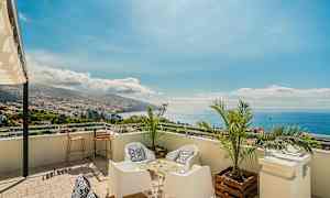 Utsikt från en terrass med sittmöbler, palmer och panoramavy över stad, berg och hav. (AI-genererad text)