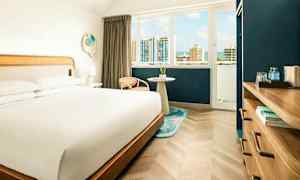 Ett hotellrum med en stor säng, ett fönster med utsikt över en stad med höghus och himmel, samt en möbel med vattenflaskor och böcker. (AI-genererad text)