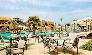 Uteservering vid en pool i en trädgårdsliknande miljö, omgiven av palmer och byggnader. (AI-genererad text)