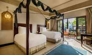 Ett sovrum med två enkelsängar, mörka trämöbler och stora glasdörrar med utsikt mot en terrass. (AI-genererad text)