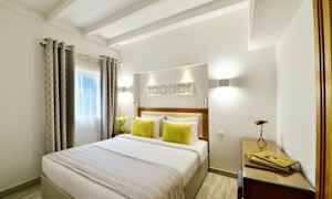 Ett ljust sovrum med en dubbelsäng, dekorerad med två gula kuddar och ett överkast. Fönstret har gardiner och det finns en arbetsyta med skrivbord och lampa. (AI-genererad text)