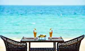 Två rottingstolar runt ett bord med två glas juice och en skål med färsk frukt på en tropisk strand. (AI-genererad text)