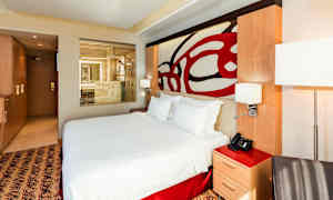 Ett modernt hotellrum med en stor säng och genomskinligt glasparti mot ett badrum. (AI-genererad text)