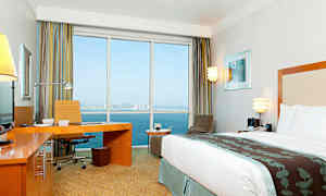 Ett modernt hotellrum med en stor säng, skrivbord och utsikt genom stora fönster mot havet och en stad i bakgrunden. (AI-genererad text)