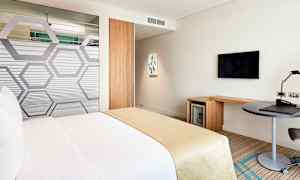 En hotellrum med en säng klädd i vita och beige färger. Rummet innehåller också ett skrivbord, en TV på väggen och ett minikylskåp. (AI-genererad text)