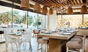 Interiör av en restaurang med bord dukade med porslin och glas samt rottinglampor hängande från taket. Utanför syns palmer. (AI-genererad text)