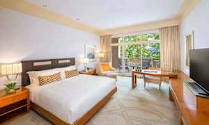 Ett hotellrum med en stor säng, en skrivbordsyta och en balkong med utsikt över grönska. (AI-genererad text)