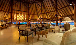 Ett lyxigt sittområde med trästolar och soffor, elegant belysning och en utsikt över havet genom öppna väggar. (AI-genererad text)