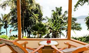 Träterrass med två stolar och ett bord med utsikt över tropiska palmer och blått hav. (AI-genererad text)