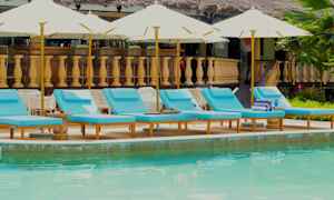 Bild på en pool med flera blå solstolar och parasoller i förgrunden, och en byggnad med träbalustrad i bakgrunden. (AI-genererad text)