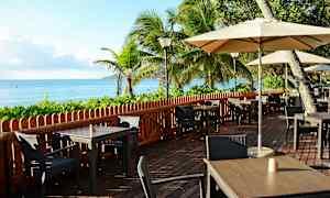 Uteservering vid havet med bord, stolar och parasoller, omgiven av grönska och palmträd. (AI-genererad text)