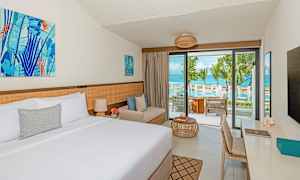 Ett hotellrum med en dubbelsäng, en sittplats och en balkong med utsikt över en pool och havet. (AI-genererad text)