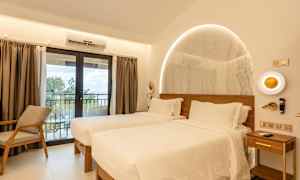 Ett hotellrum med två enkelsängar, ljus inredning och ett stort fönster med gardiner som leder till en balkong. (AI-genererad text)