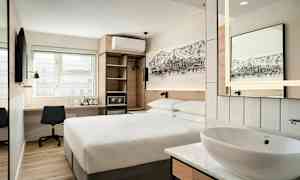 Ett modernt hotellrum med en dubbelsäng, ett skrivbord med stol, en väggmonterad TV, samt en öppen garderob och en liten tvättställ med spegel. (AI-genererad text)