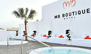 Ett hotellområde med vita solstolar och orange handdukar vid en pool, samt en palm och vita väggar. (AI-genererad text)