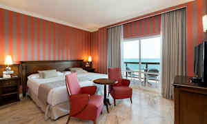 Dubbelrum med två röda fåtöljer, ett bord, havsutsikt genom en balkong. (AI-genererad text)