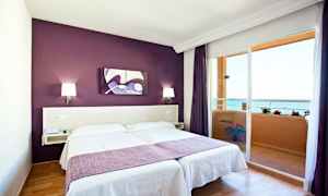 Rum med dubbelsäng, lila vägg och balkong med havsutsikt. (AI-genererad text)