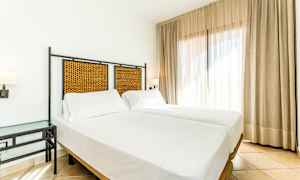 Ett ljust hotellrum med två enkelsängar, vita lakan och en flätad sänggavel. Rummet har ett sidobord och stora fönster med gardiner. (AI-genererad text)