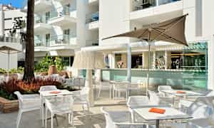 En utomhusmiljö med bord, stolar och parasoller framför en byggnad. (AI-genererad text)