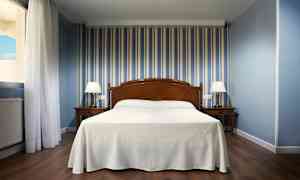 Hotellrum med säng i mitten, två sängbord med lampor och blårandig tapet. (AI-genererad text)