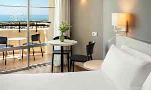 Ett hotellrum med en säng, bord och stolar nära en balkong med utsikt över havet. (AI-genererad text)