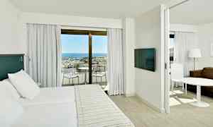 En hotellrumsinteriör med en dubbelsäng, ett separat sittområde samt en balkong med utsikt över havet och en marina. (AI-genererad text)