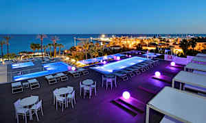 Utomhuspoolområde med utsikt över havet och en småbåtshamn, belysning i mörkret. (AI-genererad text)