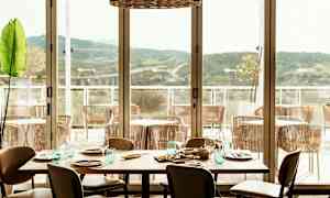 En restaurang med ett dukat bord och utsikt över en terrass med stolar och berg i fjärran. (AI-genererad text)