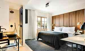 Ett hotellrum med en stor säng klädd i mörkgrått sängöverkast, en träpanel som sänggavel, en arbetsplats med bord och stol, samt ett synligt badrum med dusch bakom en glasdörr. (AI-genererad text)