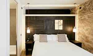 Ett hotellrum med en dubbelsäng, två hängande lampor och en inbyggd ljuspanel på träväggen bakom sängen. (AI-genererad text)