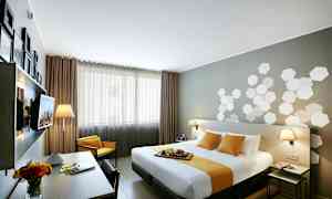 Ett modernt hotellrum med en dubbelsäng täckt av vita och gula textilier. I rummet finns ett skrivbord med en blomvas och lampor på nattduksborden. (AI-genererad text)