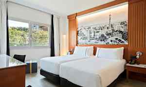 Ett hotellrum med två enkelsängar, ett skrivbord, en fönstervy mot grönska och en vägg med en stor svartvit bild som dekor. (AI-genererad text)